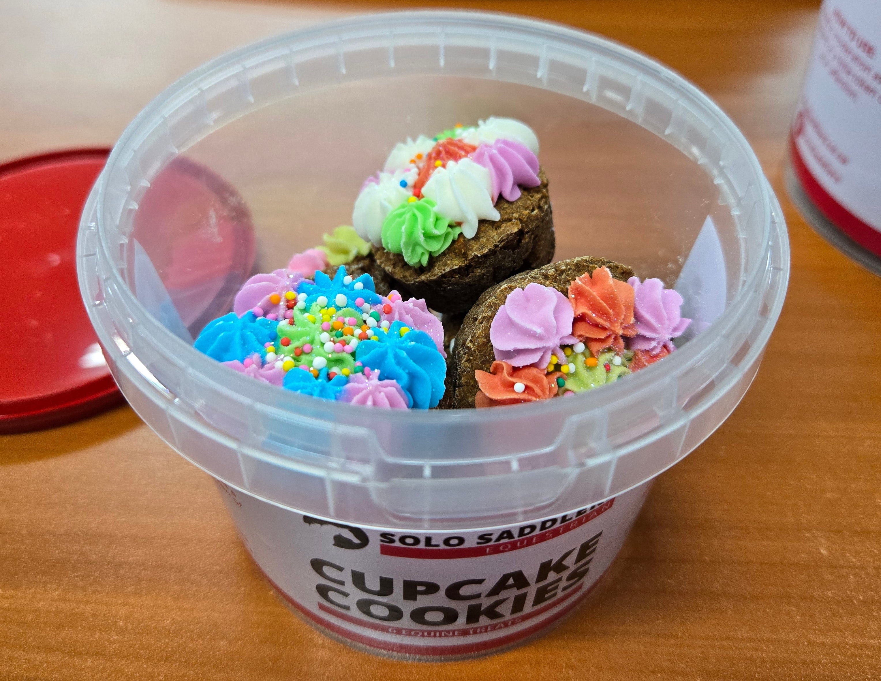 Container of cupcake cookies with colorful sprinkles on a wooden desk.