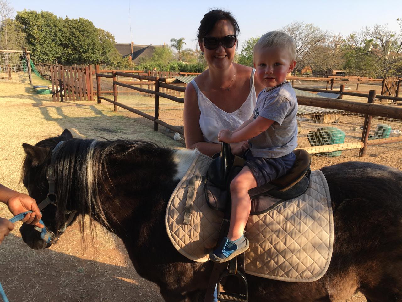 A woman wearing sunglasses and a white tank top sits on a black pony with a child riding on her lap. The pony has a quilted beige saddle pad, and the photo is taken outdoors in a sunny paddock with wooden fences and trees in the background.