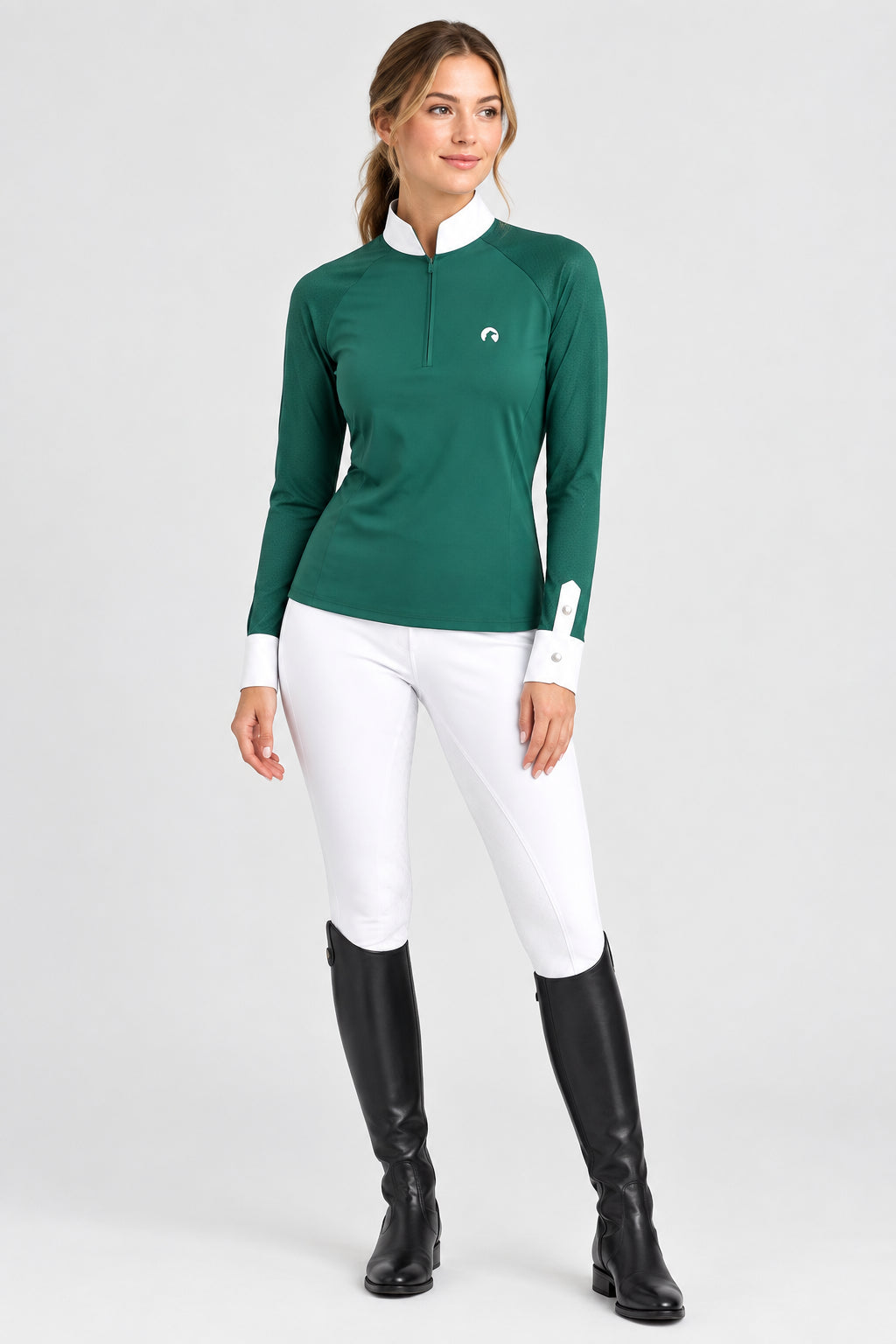 A woman modeling a dark green long-sleeve equestrian quarter-zip shirt with a white collar and white side panels, paired with white breeches and black riding boots, against a light gray studio background.